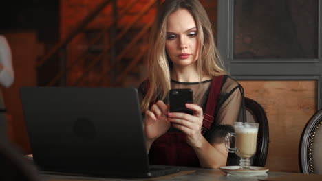 Young-woman-sitting-in-coffee-shop-at-wooden-table,-drinking-coffee-and-using-smartphone.On-table-is-laptop.-Girl-browsing-internet,-chatting,-blogging.-Female-holding-phone-and-looking-on-his-screen.